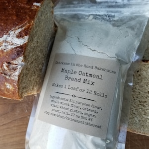 May include: A clear plastic bag filled with a light gray bread mix. The bag has a label that reads "Chickens in the Road Bakehouse Maple Oatmeal Bread Mix Makes 1 Loaf or 12 Rolls Ingredients: All purpose flour, whole wheat flour, oatmeal, vital wheat gluten, sugar, yeast, salt. 17 oz Net Wt etsy.com/shop/chickensintheroad". Two slices of bread are visible on either side of the bag.