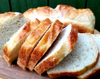 May include: Sliced loaf of golden-brown bread, with a soft, white interior. The bread is on a wooden cutting board, with a whole loaf in the background. The crust has a shiny, baked appearance.
