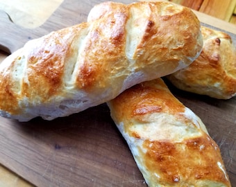 May include: Three golden brown baguettes on a wooden cutting board. The baguettes are slightly flattened and have a crispy crust.