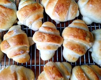 May include: A close-up of a wire rack with a dozen golden brown crescent rolls. The rolls are arranged in a staggered pattern, with some rolls overlapping.