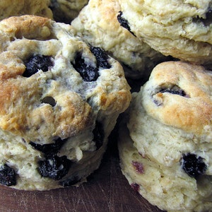 May include: A close-up of a stack of freshly baked blueberry biscuits. The biscuits are golden brown and have a slightly crispy exterior. The blueberries are visible throughout the biscuits, adding a touch of sweetness and color.