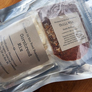 May include: Two clear plastic bags containing a cornbread mix and a chili mix. The cornbread mix bag has a brown label with the text "Chickens in the Road Bakehouse Cornbread Mix" and a list of ingredients. The chili mix bag has a brown label with the text "Chickens in the Road Bakehouse Chili Mix Heat: Medium" and a list of ingredients.