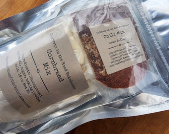May include: Two clear plastic bags containing a cornbread mix and a chili mix. The cornbread mix bag has a brown label with the text "Chickens in the Road Bakehouse Cornbread Mix" and a list of ingredients. The chili mix bag has a brown label with the text "Chickens in the Road Bakehouse Chili Mix Heat: Medium" and a list of ingredients.