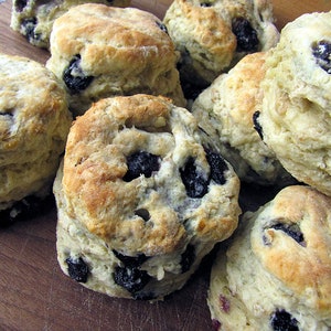 May include: A close-up of a batch of freshly baked blueberry biscuits. The biscuits are golden brown and have a slightly crispy exterior. The blueberries are visible throughout the biscuits, adding a touch of sweetness and color.