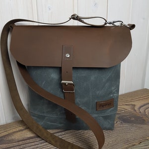 Może przedstawiać: Szara torba kurierska z płótna z brązową skórzaną klapą i paskiem. Torba ma zapięcie na klamrę i małą prostokątną etykietę z napisem "Papivert". Torba leży na drewnianej powierzchni.