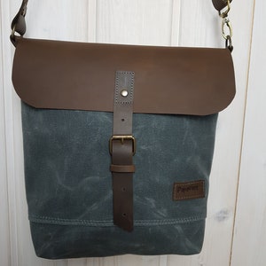 Może przedstawiać: Szara torba listonoszka z płótna z brązową klapą i paskiem ze skóry. Torba ma mosiężną klamrę i małą skórzaną naszywkę z napisem "Prevert".