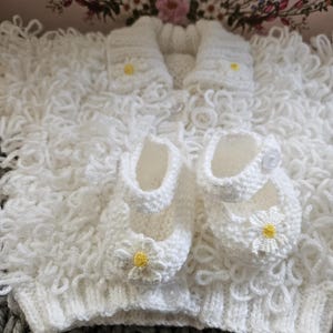 May include: A white knitted baby set, comprising a sweater, booties, and mittens, each decorated with a small yellow and white daisy. The set is presented on a textured white surface, with a grey and white knitted blanket in the background.