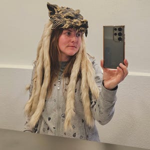 Könnte beinhalten: Ein detaillierter, bronzefarbener Wolfskopfschmuck mit langem, hellfarbenem Fell. Der Kopfschmuck weist aufwendige Details des Wolfsgesichts auf und wird mit einem grauen Kapuzenpullover mit Reißverschluss getragen. Das Bild ist ein Selfie vor einem Spiegel.