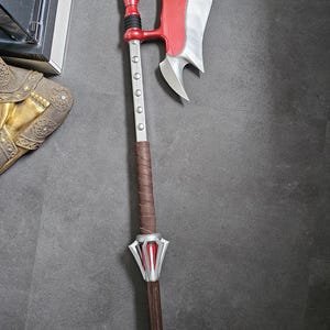 Può includere: Un'ascia da battaglia rossa e argento con un lungo manico marrone. La testa dell'ascia è argentata con un accento rosso e il manico è avvolto in pelle marrone. L'ascia ha un'estremità appuntita.