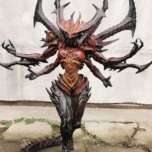 May include: A detailed sculpture of a demonic figure with multiple arms and a tail. The figure is predominantly red, black, and orange, with sharp, spiky features and a menacing expression. The sculpture appears to be a fantasy or gaming-themed collectible.