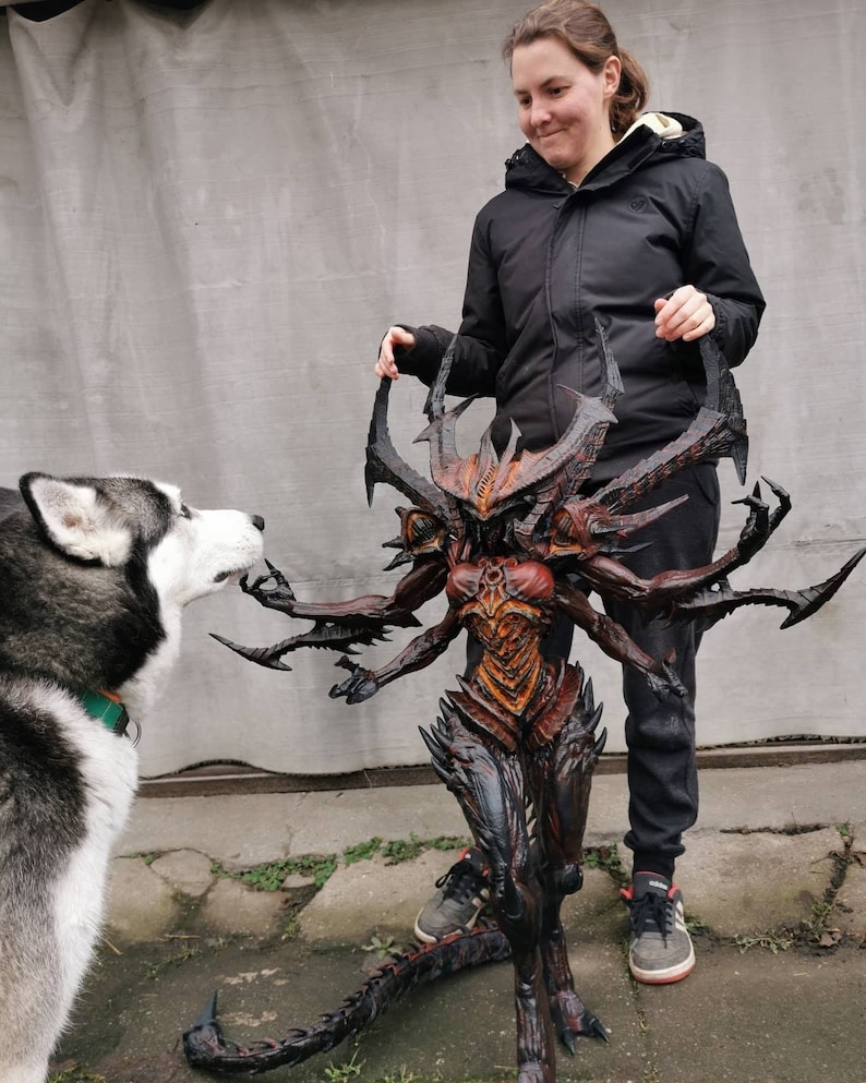 May include: A detailed sculpture of a demonic figure, predominantly dark in colour, with multiple arms and a long tail. The figure has a red and orange torso, with black and grey accents. The sculpture is held by a person.