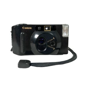 Puede incluir: Cámara Canon Sure Shot negra con flash incorporado y tapa de lente. La cámara tiene una correa de muñeca negra adjunta. La palabra "Canon" está impresa en dorado en la parte superior de la cámara.