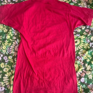 May include: A solid red t-shirt with short sleeves. The shirt is made of a soft material and has a crew neck. The shirt is displayed on a floral patterned fabric.
