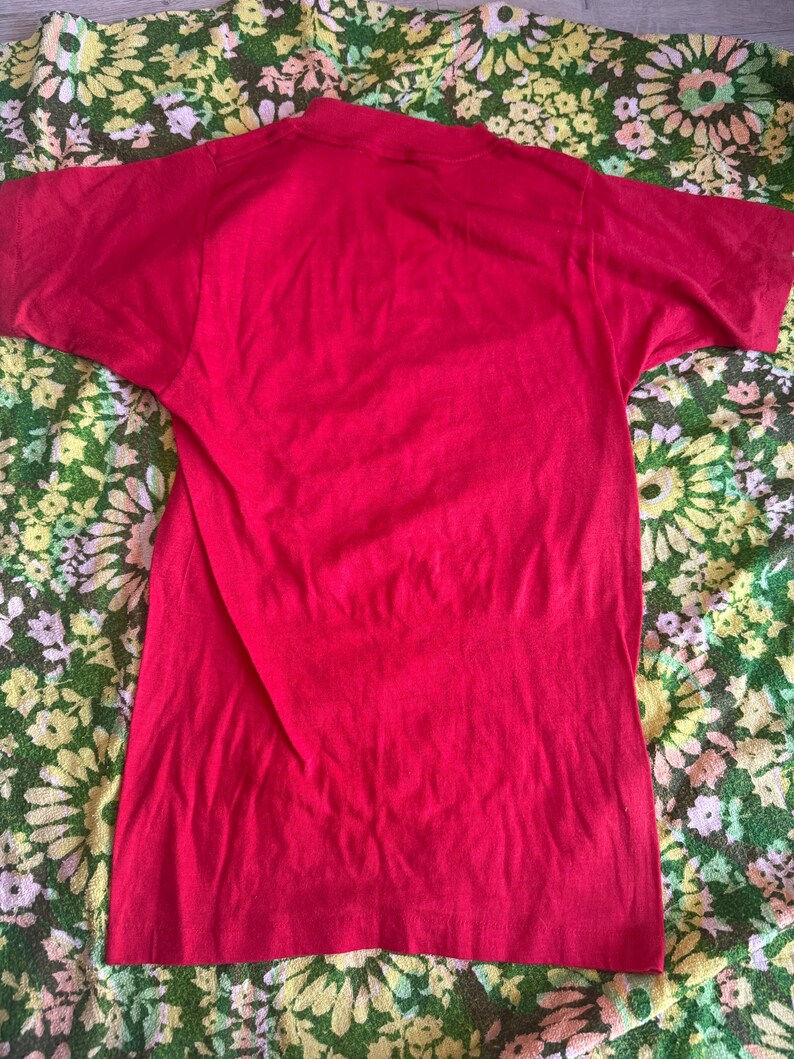 May include: A solid red t-shirt with short sleeves. The shirt is made of a soft material and has a crew neck. The shirt is displayed on a floral patterned fabric.