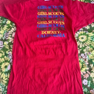 May include: A red t-shirt with the text "GIRL SCOUTS" stacked vertically in multiple colors. Below the text is "DOWNEY, CALIFORNIA" in blue. The shirt is made of a soft material and has short sleeves.