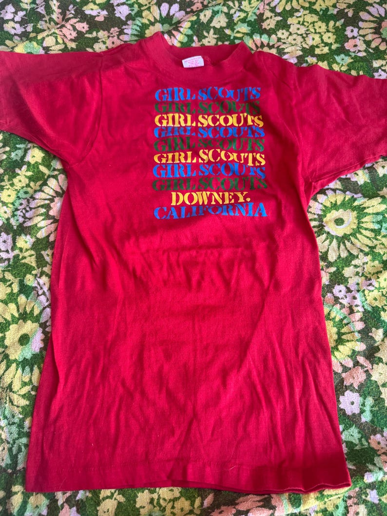 May include: A red t-shirt with the text "GIRL SCOUTS" stacked vertically in multiple colors. Below the text is "DOWNEY, CALIFORNIA" in blue. The shirt is made of a soft material and has short sleeves.
