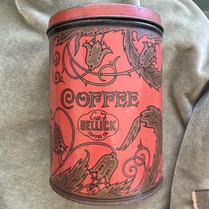 May include: A cylindrical metal coffee canister with a red background and dark brown floral illustrations. The text reads COFFEE and Geo. F. HELLICK COFFEE CO. within a central oval emblem. The container features a matching red lid.