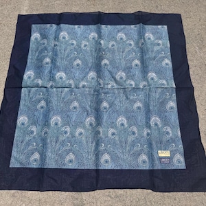 May include: A square scarf featuring a central panel with a blue and white peacock feather pattern, surrounded by a solid dark navy blue border. A small rectangular tag with the text Liberty of London is attached to the lower right corner.