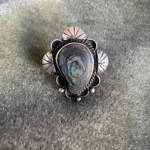 May include: A silver-toned brooch with an oval, iridescent center stone. The stone is surrounded by a decorative silver frame with a scalloped edge and small, stylized fan shapes. The brooch is set against a green velvet background.