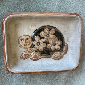 May include: A rectangular ceramic dish with a beige textured base and a brown rim. The center features a raised relief of a turtle with a dark brown shell decorated with three light brown flowers, each containing a small blue center dot.