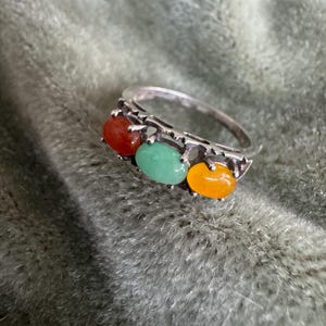 May include: A silver ring featuring three oval-shaped cabochon stones set in a row. The stones are colored red, light green, and orange. The metal band has a decorative, notched edge design that holds each of the three smooth, polished stones.