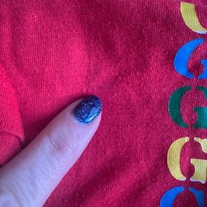 May include: Close-up of a red fabric with the word "G" in blue, green, yellow, and blue. The fabric is slightly textured. A finger with blue glitter nail polish is touching the fabric.