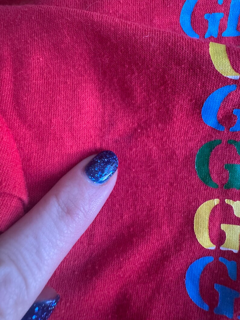 May include: Close-up of a red fabric with the word "G" in blue, green, yellow, and blue. The fabric is slightly textured. A finger with blue glitter nail polish is touching the fabric.