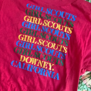 May include: A red t-shirt featuring the words "GIRL SCOUTS" in a repeating pattern of blue, green, and yellow. The text "DOWNEY, CALIFORNIA" is printed at the bottom in blue. The shirt has a crew neck.