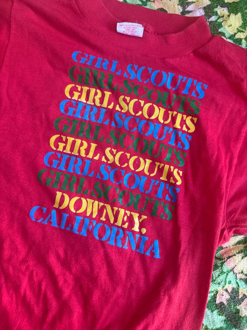 May include: A red t-shirt featuring the words "GIRL SCOUTS" in a repeating pattern of blue, green, and yellow. The text "DOWNEY, CALIFORNIA" is printed at the bottom in blue. The shirt has a crew neck.