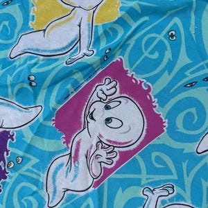 Op de afbeelding: Een stof met een herhaald patroon met het stripfiguur Casper het Vriendelijke Spook. Casper is wit met blauwe accenten, tegen een turquoise achtergrond met wervelende ontwerpen en accenten van geel en paars.