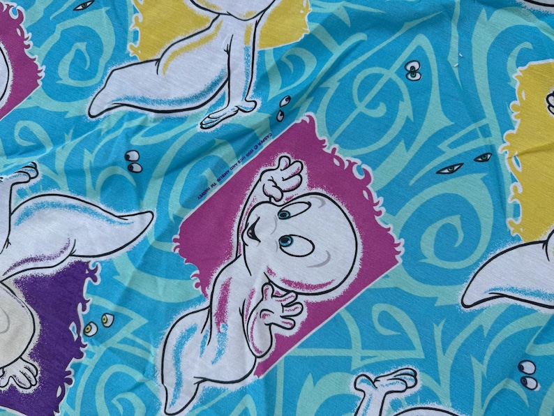 Op de afbeelding: Een stof met een herhaald patroon met het stripfiguur Casper het Vriendelijke Spook. Casper is wit met blauwe accenten, tegen een turquoise achtergrond met wervelende ontwerpen en accenten van geel en paars.