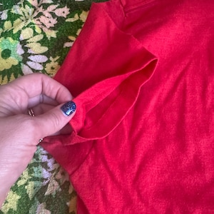 May include: Close-up of a bright red t-shirt sleeve. The sleeve is folded over, revealing the inside seam. The fabric appears to be a soft, knit material. The background is a floral pattern in green, yellow, and white.