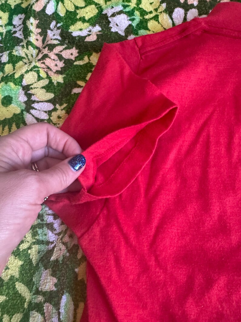 May include: Close-up of a bright red t-shirt sleeve. The sleeve is folded over, revealing the inside seam. The fabric appears to be a soft, knit material. The background is a floral pattern in green, yellow, and white.