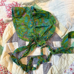 May include: A green and blue paisley patterned bonnet with a gathered ruffle around the crown and long ties. The bonnet is lying on a patchwork quilt.
