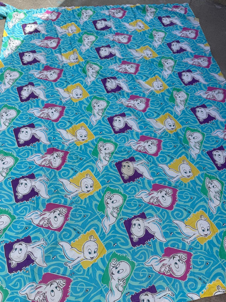 Op de afbeelding: Een lichtblauw laken met een herhaald patroon van het stripfiguur Casper het vriendelijke spook. Elke Casper staat in een kleurrijk vierkant, met een golvende blauwe achtergrond.