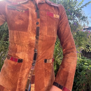 May include: A long-sleeved, button-down jacket in a warm, textured brown tone. The garment features a pointed collar, two chest pockets, and two lower pockets, all accented with decorative strips of patterned fabric in red, black, and gold.