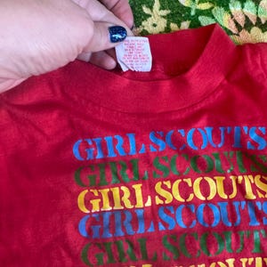 May include: A red t-shirt with the words "GIRL SCOUTS" printed in blue, green, yellow, and blue. The shirt has a crew neck and a tag is visible at the neckline.