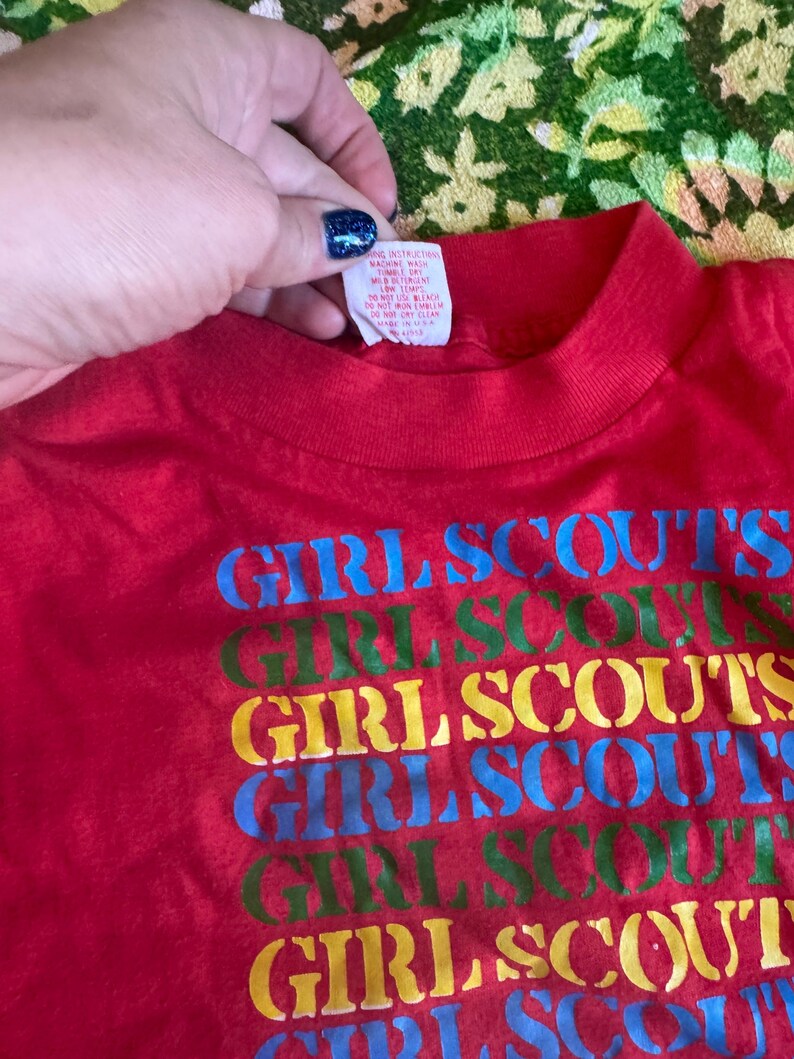 May include: A red t-shirt with the words "GIRL SCOUTS" printed in blue, green, yellow, and blue. The shirt has a crew neck and a tag is visible at the neckline.