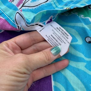 Op de afbeelding: Een close-up van een kleurrijke stof met een bloemenpatroon in blauw, paars en wit. Een label van "THE BIBS COMPANY" is zichtbaar, met de herkomst en samenstelling van de stof. De stof is gemaakt van 70% polyester en 30% katoen.