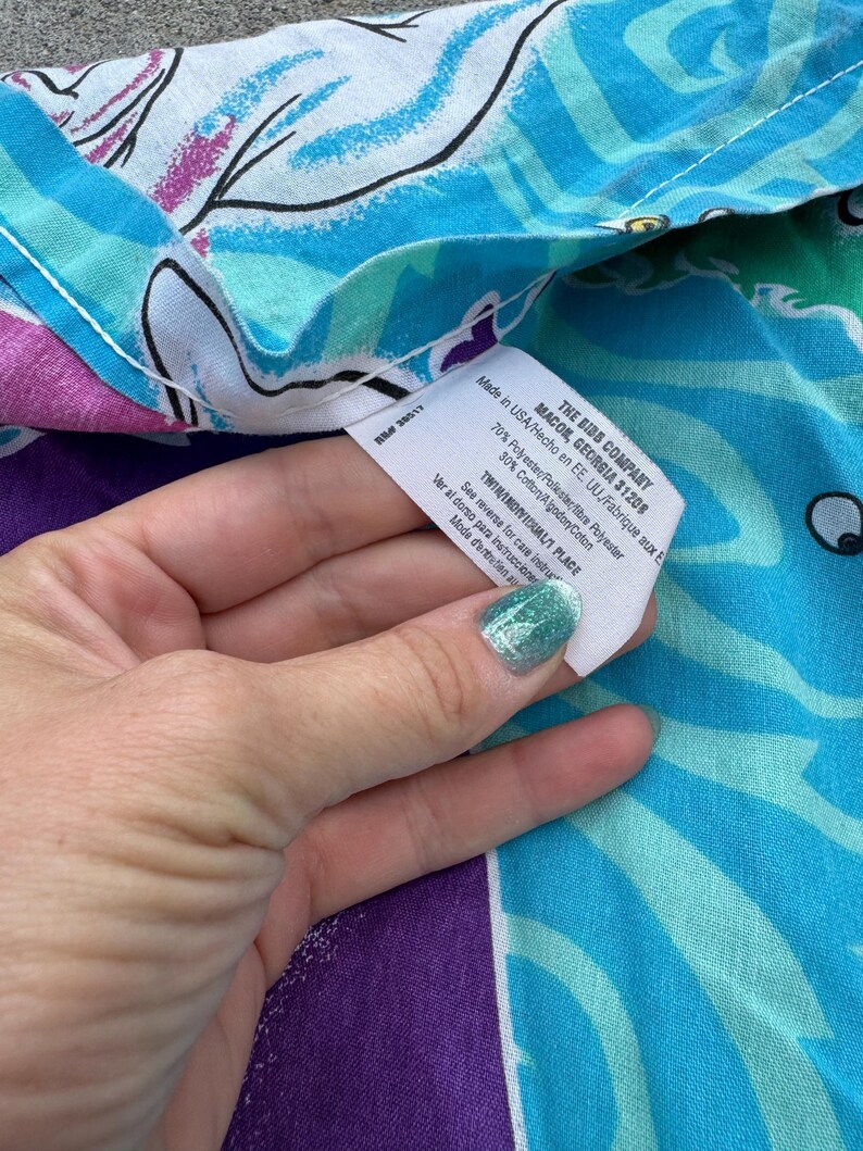 Op de afbeelding: Een close-up van een kleurrijke stof met een bloemenpatroon in blauw, paars en wit. Een label van "THE BIBS COMPANY" is zichtbaar, met de herkomst en samenstelling van de stof. De stof is gemaakt van 70% polyester en 30% katoen.