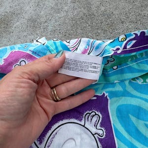 Op de afbeelding: Een gedeelte van een Casper the Friendly Ghost-stof met patronen in tinten blauw, paars en wit. De stof toont het gezicht van Casper en andere cartoon-elementen. Een hand houdt een label met wasvoorschriften vast.