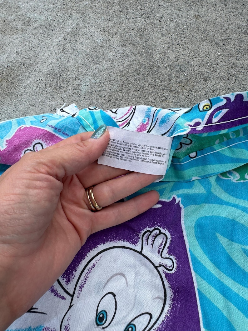 Op de afbeelding: Een gedeelte van een Casper the Friendly Ghost-stof met patronen in tinten blauw, paars en wit. De stof toont het gezicht van Casper en andere cartoon-elementen. Een hand houdt een label met wasvoorschriften vast.