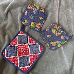 May include: Three quilted pot holders. Two are square with a denim-like blue fabric and a floral pattern. The third is a square with a red bandana pattern and denim-like blue squares. All have blue trim.