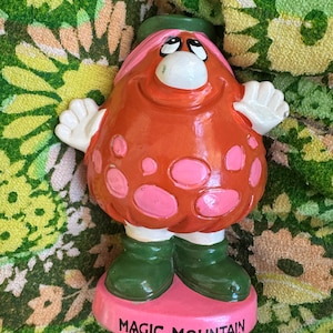 May include: A ceramic figurine of a smiling, cartoonish character with a round, orange body and pink spots. It has white hands, green boots, and a green hat. The figurine is on a pink base with the words "MAGIC MOUNTAIN" in white.