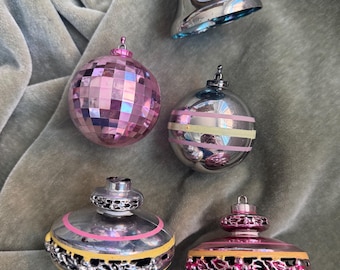 May include: A collection of five decorative ornaments including a silver bell with pink and yellow stripes, a pink disco ball sphere, a silver sphere with pink and yellow stripes, and two ornate silver and pink finial-shaped ornaments with cutouts.