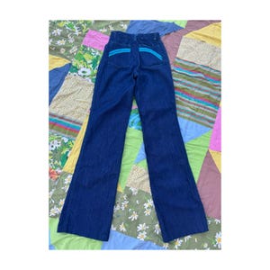 May include: Dark blue denim jeans with a high waist and flared legs. The back pockets feature a turquoise accent. The jeans are laid flat on a colorful patchwork quilt.