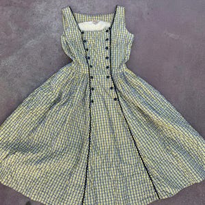 May include: A sleeveless dress featuring a yellow and blue checkered pattern. The bodice has a double-breasted design with two vertical rows of dark buttons and black rickrack trim. The skirt is full and pleated, extending from the waistline.