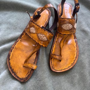May include: A pair of mustard yellow leather sandals featuring a toe loop, an adjustable ankle strap with a black buckle, and a decorative band across the instep with woven tan fabric inserts. The edges are finished with dark brown stitching.