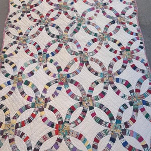 May include: A double wedding ring pattern quilt featuring interlocking rings made from various multicolored fabric scraps. The quilt has a white background with diagonal quilted stitching throughout and a thin red and white checkered border.