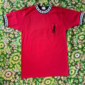 May include: Red short-sleeved t-shirt with a black and white striped collar and a small pocket on the left side. The pocket has a black and white graphic of a lighthouse and the text "Yarmouth".